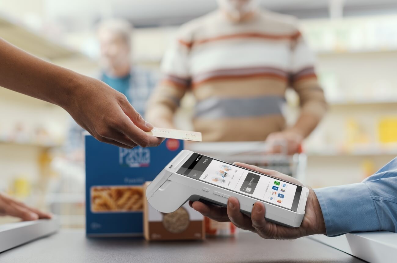 Cashier holding a Valor handheld terminal while customer taps their card at a grocery checkout